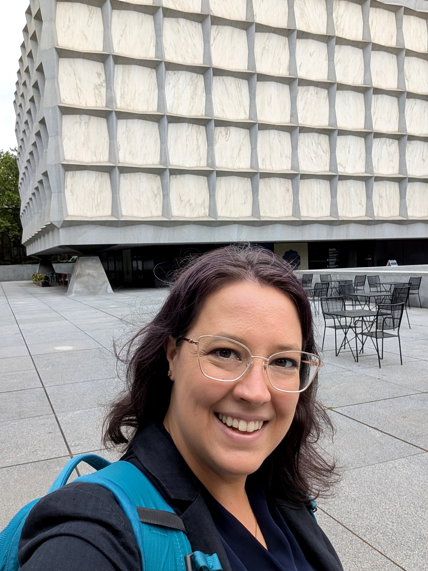 Selfie photo of a woman with dark brown shoulder length hair and glasses, who is smiling and standing in front of a square cement-colored buidling with no windows.