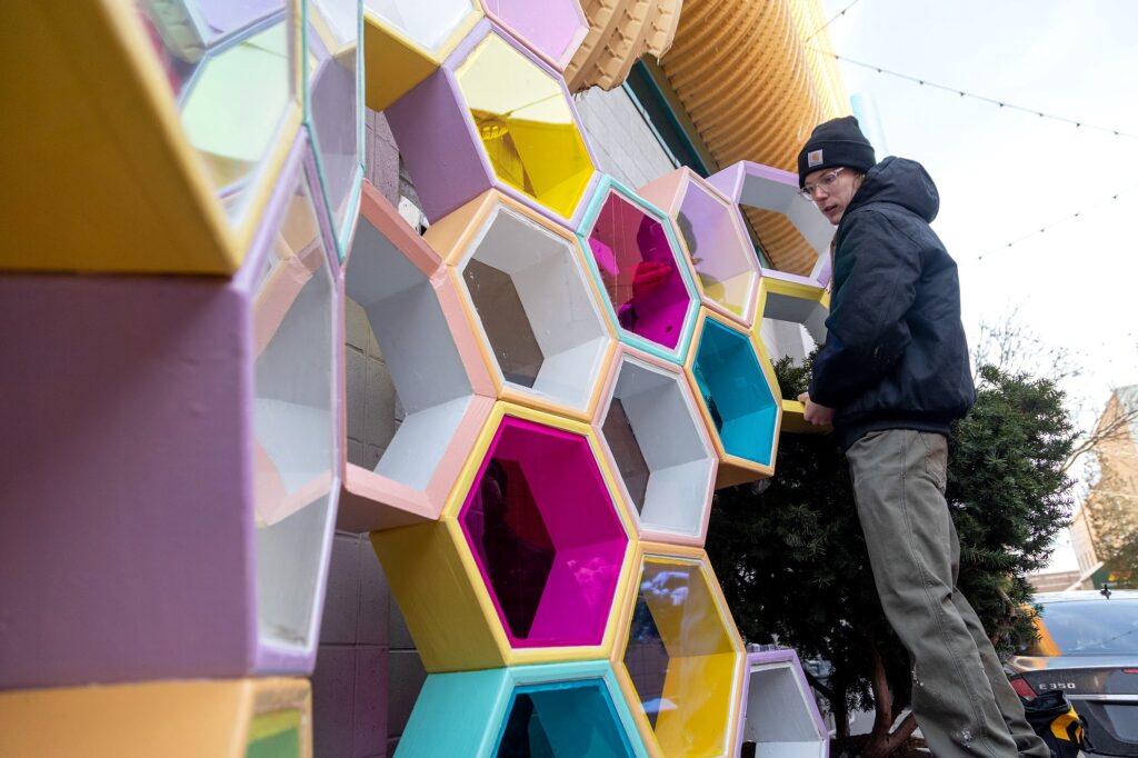 A person in a winter jacket and hat works beside the installed sculpture, reaching into one of the colorful hexagonal modules. The structure’s translucent panels reflect the surrounding area as it rises vertically along the wall and into a cluster of evergreen branches.
