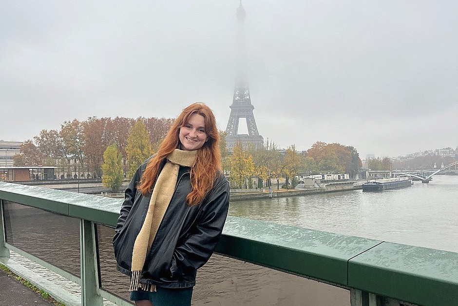 A young woman with long red hair stands on a bridge over the Seine River, wearing a black jacket and a beige scarf. Fog partially obscures the Eiffel Tower in the background, and autumn trees line the riverbank.