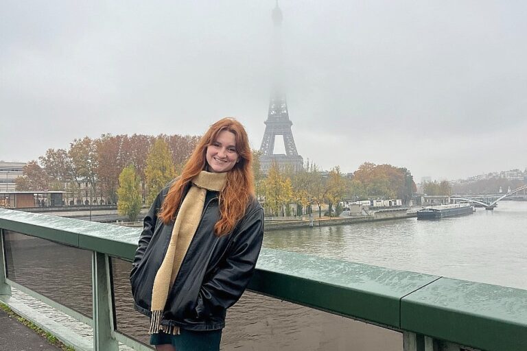 A young adult with long red hair stands on a bridge over the Seine River, wearing a black jacket and a beige scarf. Fog partially obscures the Eiffel Tower in the background, and autumn trees line the riverbank.
