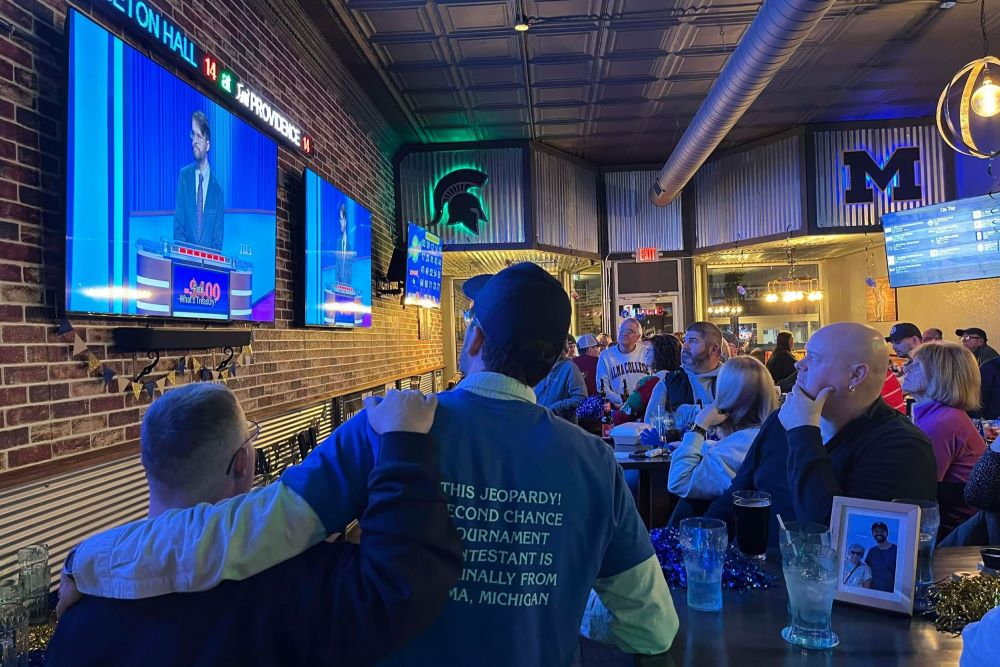 Two people with their arms around each other watch the “Jeopardy!” game show on multiple TV screens in a restaurant setting.