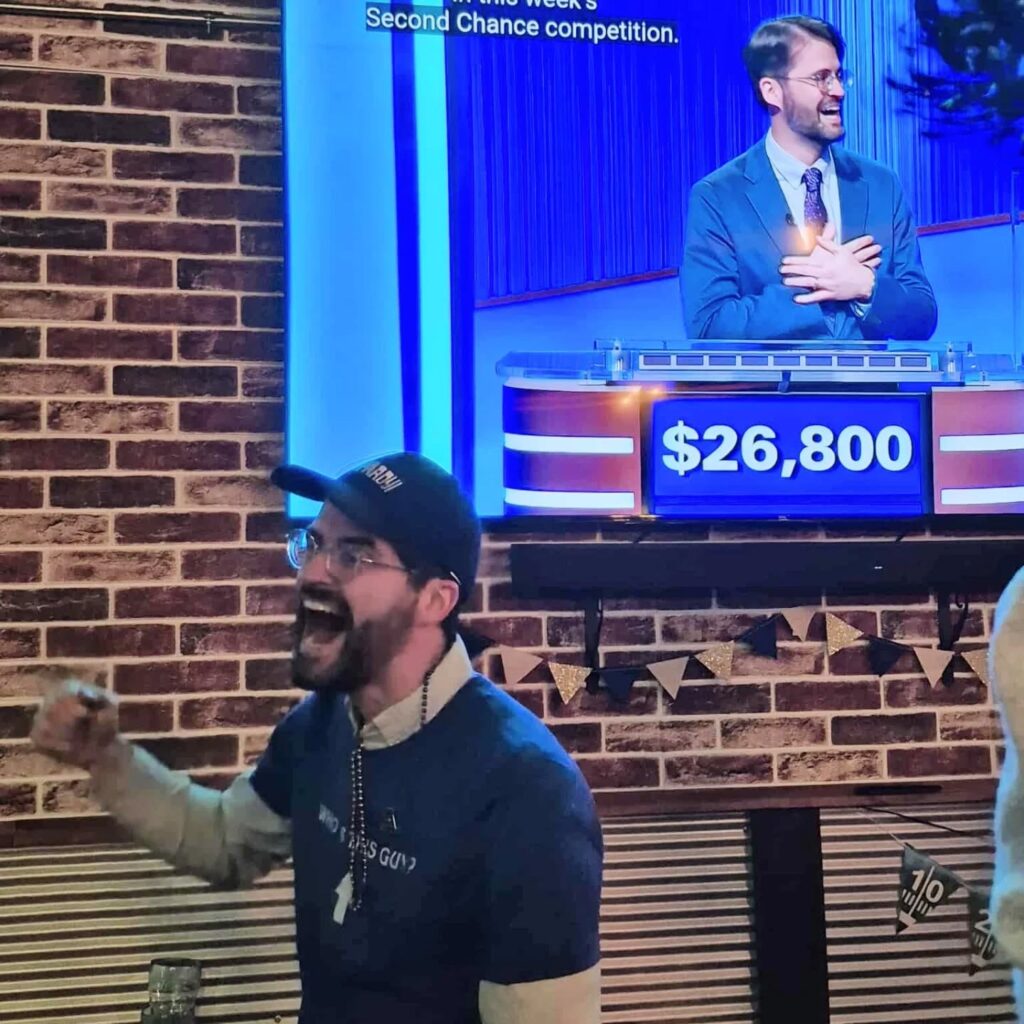 Peter Johnson cheers and his fist up. Behind him is a TV screen with the image of him winning on the “Jeopardy!” game show.