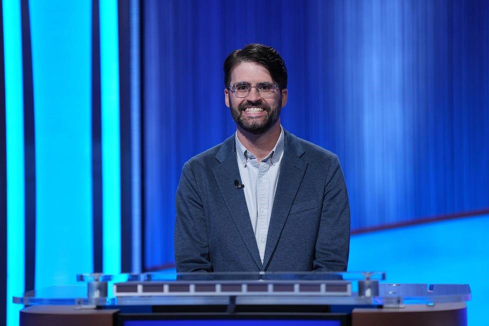A person in a blazer behind a podium on the "Jeopardy!" TV game show set. A blue curtain is in the background. The person is MSU Film Studies faculty member Peter Johnston.