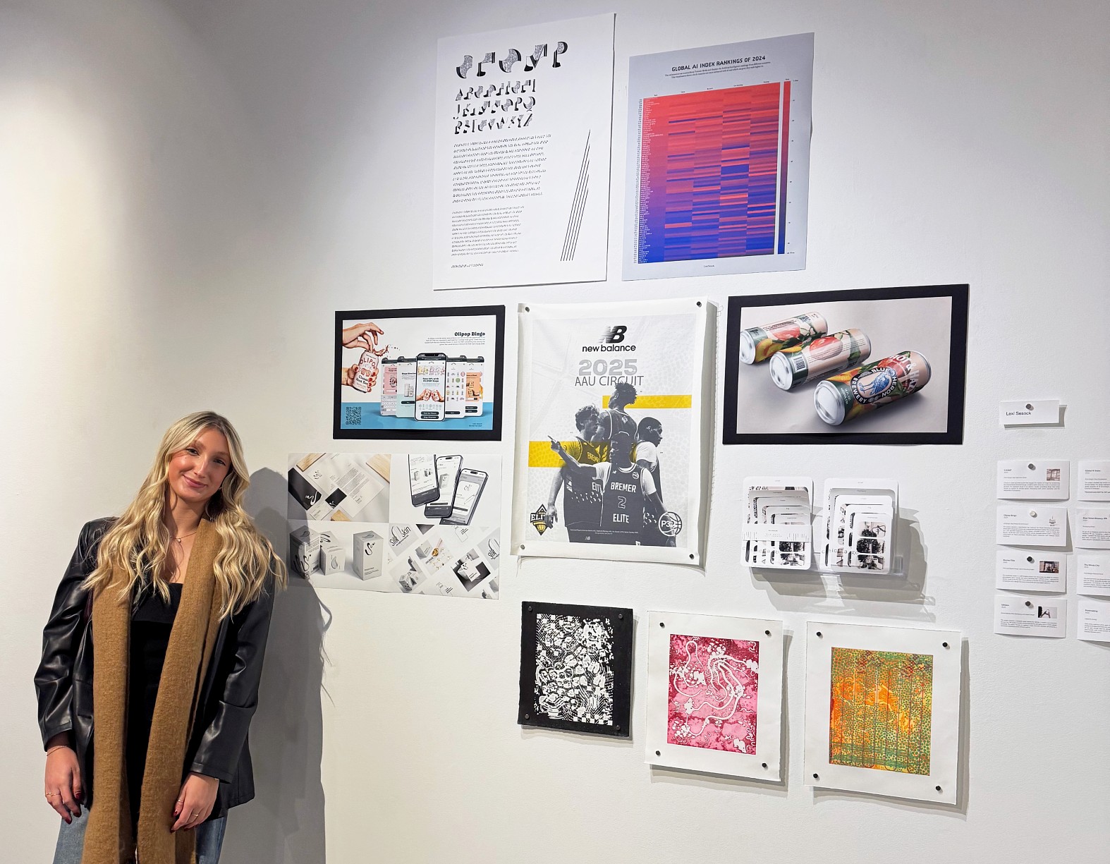 A young blonde woman stands in a gallery space next to a wall display of her creative work. She is wearing a black leather jacket, dark top, denim pants, and a long brown scarf. The wall display includes graphic design posters, framed screen-based visual concepts, photography of consumer packaging, and framed abstract prints.