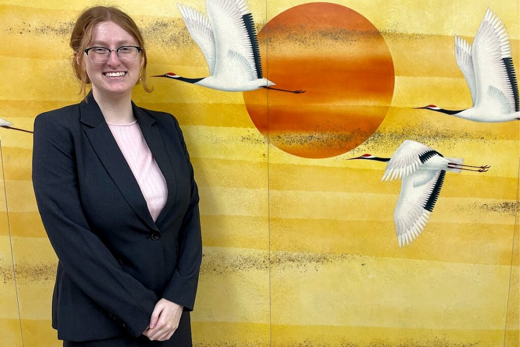 A person wearing glasses, a black blazer, and a light pink top smiles while standing in front of a yellow mural depicting white cranes flying across a large orange sun.