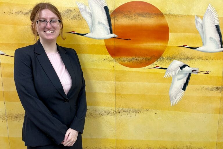 A person wearing glasses, a black blazer, and a light pink top smiles while standing in front of a yellow mural depicting white cranes flying across a large orange sun.