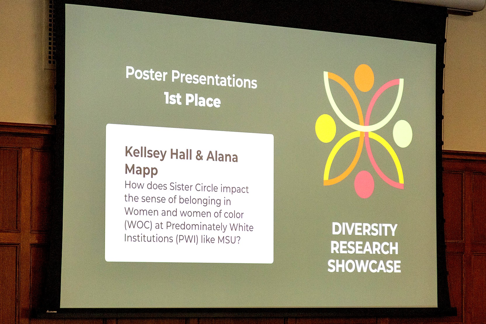A projected slide reads “Poster Presentations, 1st Place” and lists Kellsey Hall and Alana Mapp with the project title about Sister Circle and sense of belonging at predominantly white institutions, alongside the Diversity Research Showcase logo.