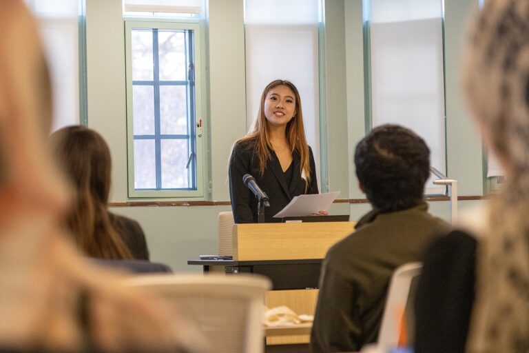 Rachel Zhai stands at a podium holding notes while delivering an oral presentation to an audience seated in a lecture-style room at the Diversity Research Showcase.