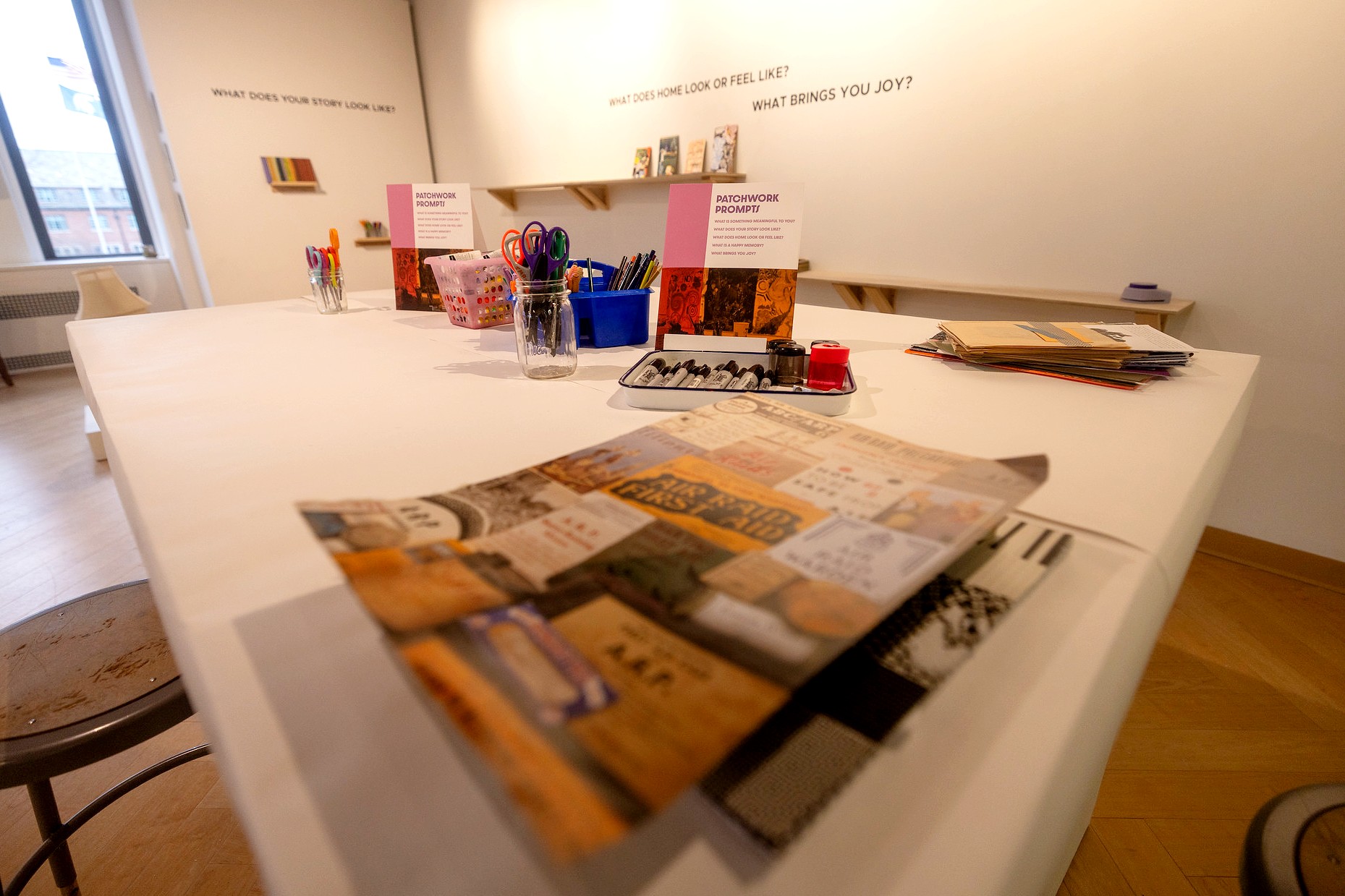 A high-angle view of a long, white communal table inside the MSU Union Art Gallery. The table is stocked with jars of scissors, bins of markers, and stacks of paper. "Patchwork Prompts" instruction cards are placed alongside various collage materials, including vintage pamphlets. In the background, vinyl lettering on the gallery wall reads "WHAT DOES HOME LOOK OR FEEL LIKE?" and "WHAT BRINGS YOU JOY?"