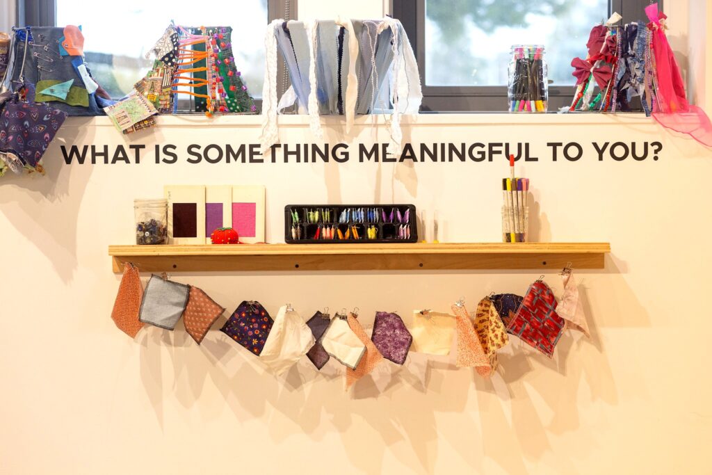 A close-up of an interactive station in the gallery. Black vinyl lettering on the wall asks, "WHAT IS SOMETHING MEANINGFUL TO YOU?" Below the text, a wooden shelf holds jars of buttons, embroidery floss, and pincushions. A string of colorful fabric patches is pinned beneath the shelf, and several decorated lampshades sit on the ledge above.