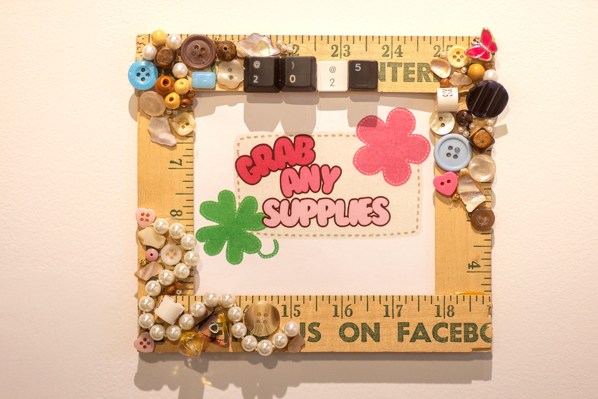 A square art piece constructed from a wooden yardstick frame. The frame is heavily embellished with assorted buttons, pearls, typewriter keys reading "2025," and a pink butterfly charm. Inside the frame, a central graphic features the text "GRAB ANY SUPPLIES" in pink and red bubble letters, accompanied by a green four-leaf clover and a pink flower.