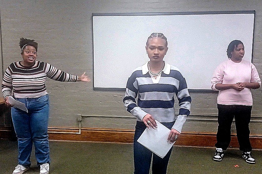 Three performers rehearse in a classroom setting, wearing casual clothing. They hold scripts while standing in front of a whiteboard on a light-colored brick wall.