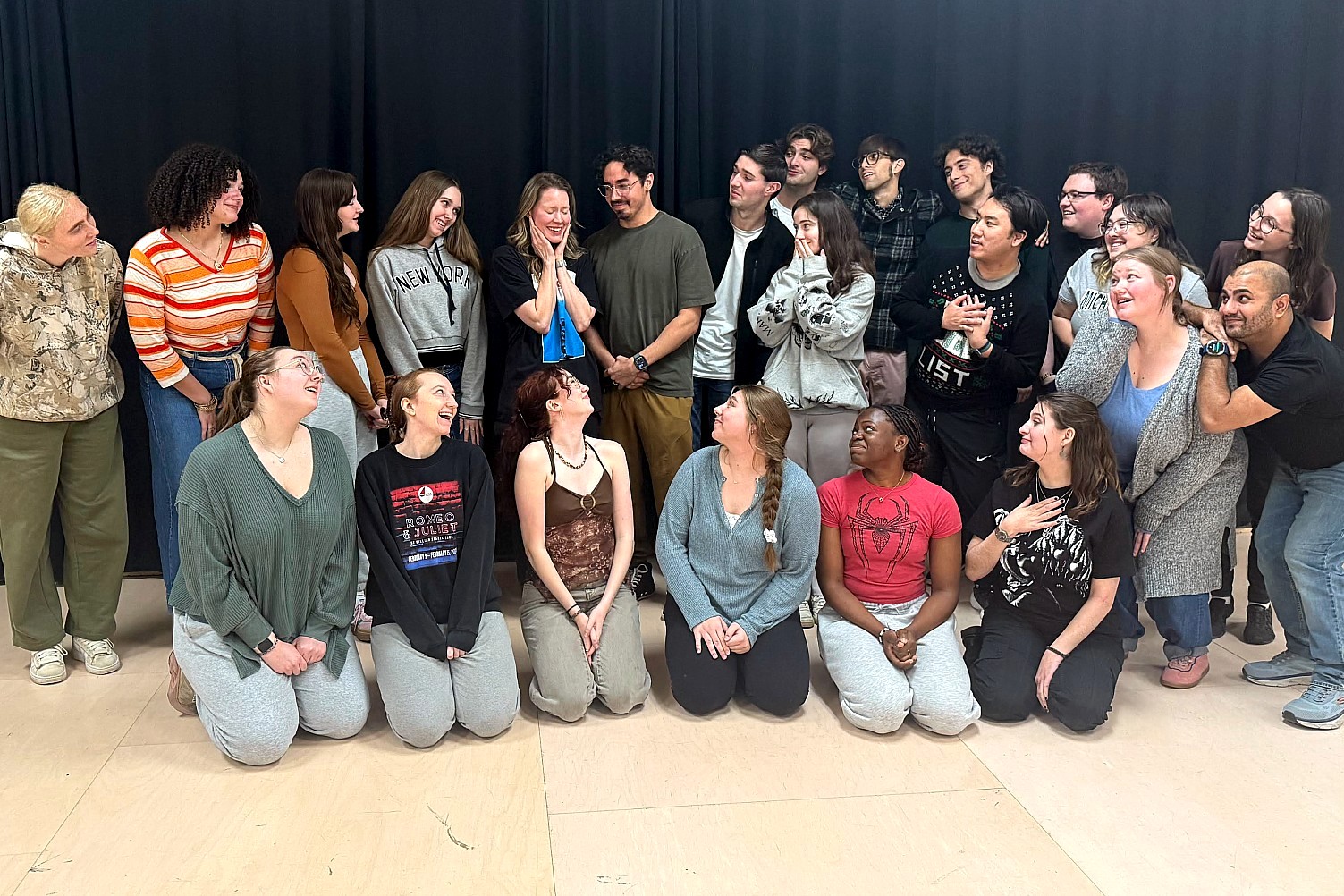 A group of approximately twenty Michigan State University theatre students and alumni posing together in a studio. They are arranged in two rows, with several individuals kneeling in the front. The group is smiling and looking at one another against a black curtain backdrop.