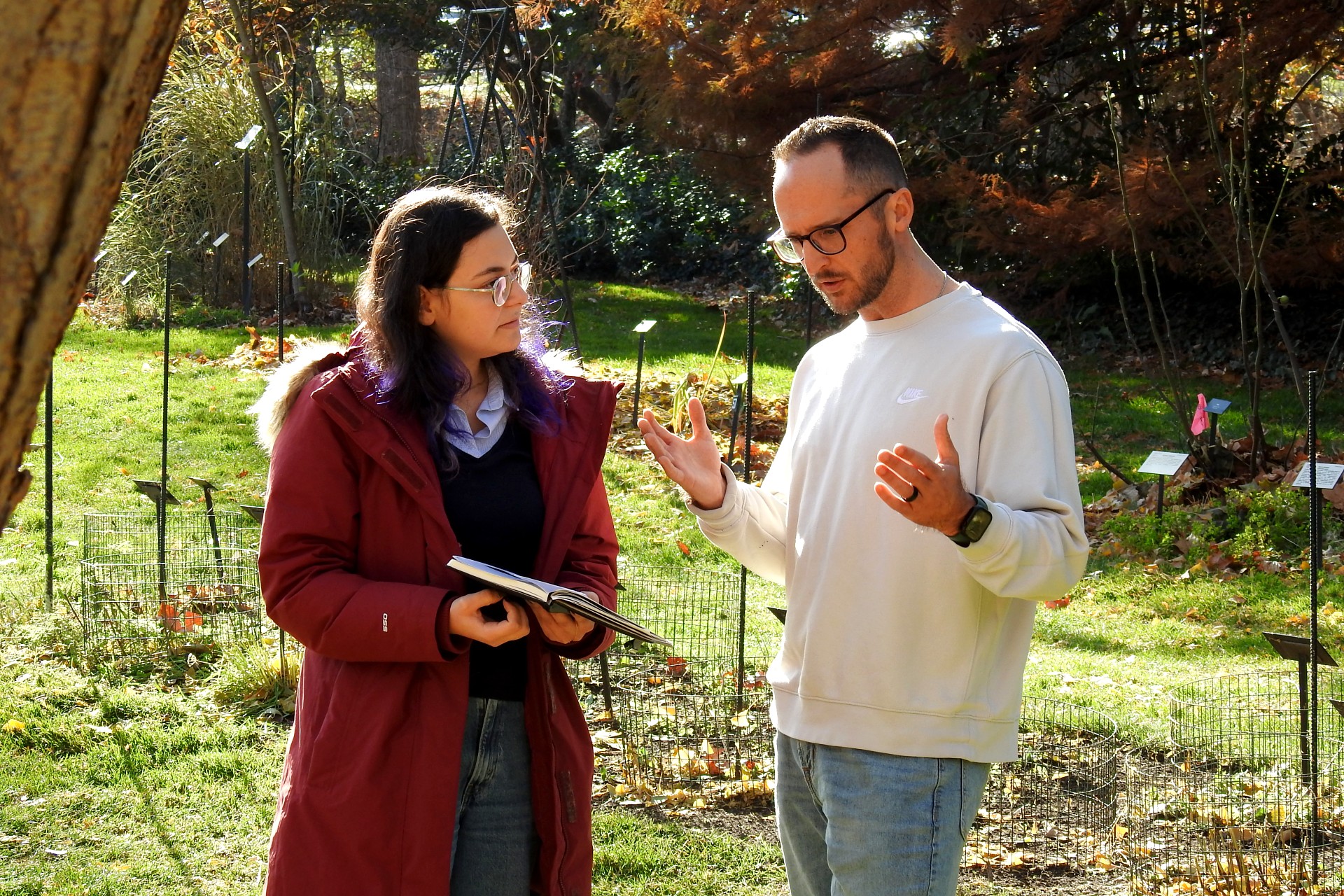 A man in a white shirt and glasses speaks with a student in a red winter coat at Beal Botanical Garden. The student holds a notebook.