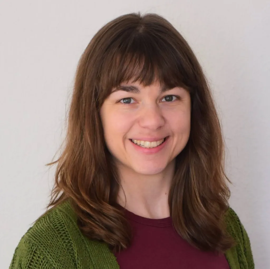 A head-and-shoulders portrait of a person with long brown hair and bangs wearing a maroon top.