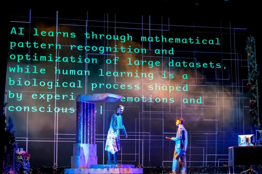 Two student performers stand on stage beneath a large projection reading, “AI learns through mathematical pattern recognition and optimization of large datasets, while human learning is a biological process shaped by experience, emotions and consciousness.” Blue and purple lighting and stage haze frame the scene, with one performer elevated on a platform and another kneeling nearby, holding a notebook.