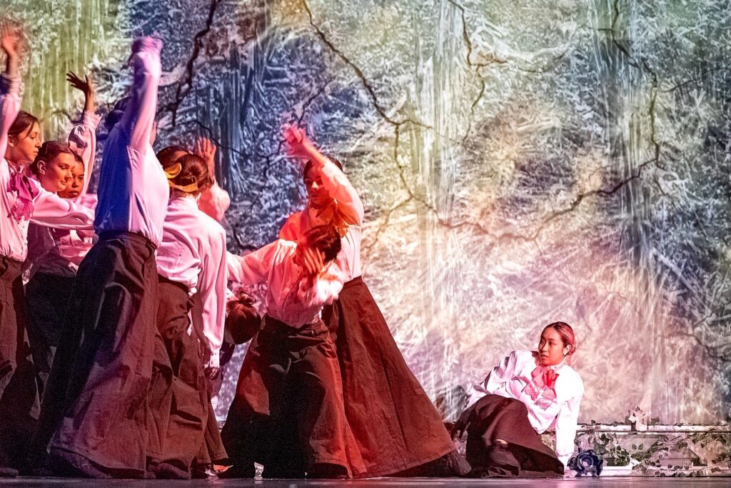 A group of student performers in long dark skirts and white blouses move in unison beneath a forest-like projection. One performer reclines on a low platform at right, watching as others raise their arms in sweeping, synchronized gestures under warm pink lighting.