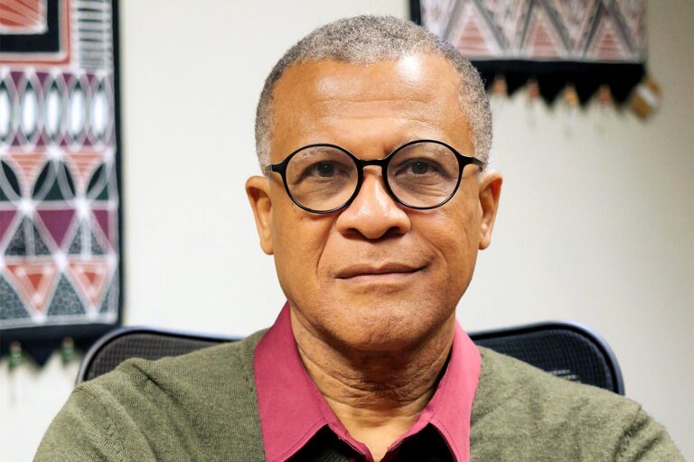 A professional headshot of Philip Effiong, Professor of Theatre Studies and Integrative Humanities. He is wearing round black-rimmed glasses, an olive green V-neck sweater over a maroon collared shirt, and a watch with a black strap.
