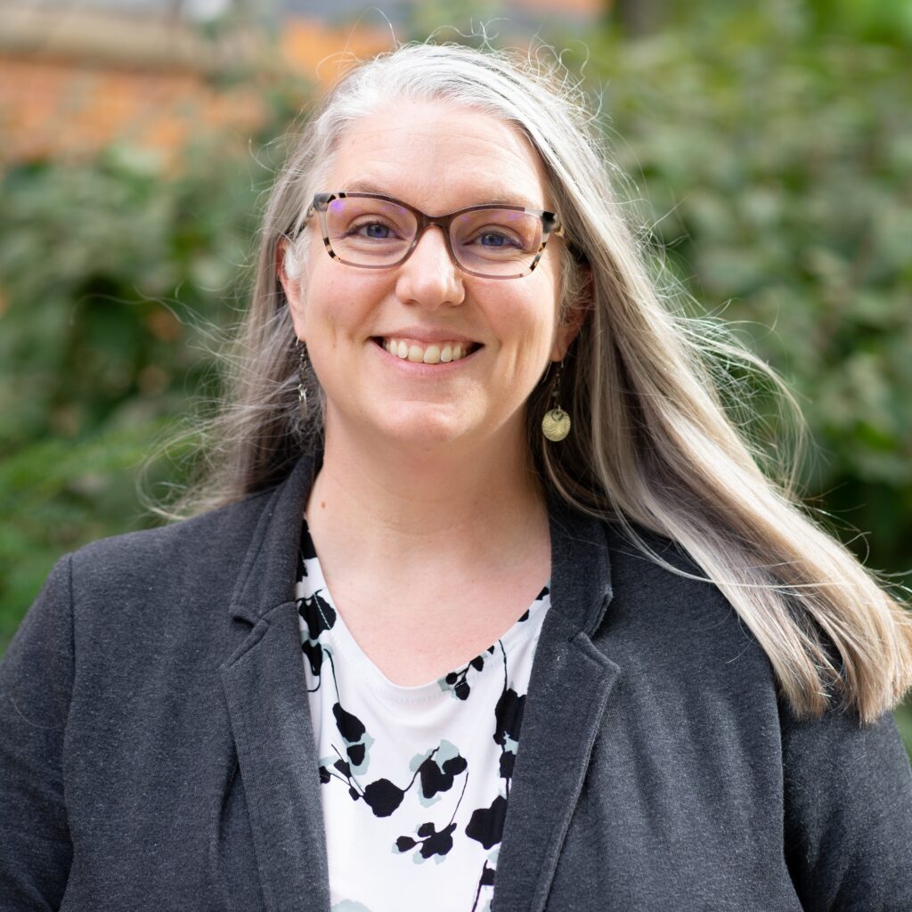 Emily Heidrich Uebel, wearing glasses and a dark cardigan over a white and black patterned top, smiling at the camera with a brick building and green foliage behind her.