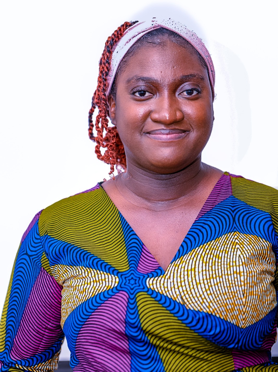 Portrait of a woman facing the camera against a white background, wearing a colorful patterned V-neck dress and a light headband, with braided hair pulled back and a slight smile.