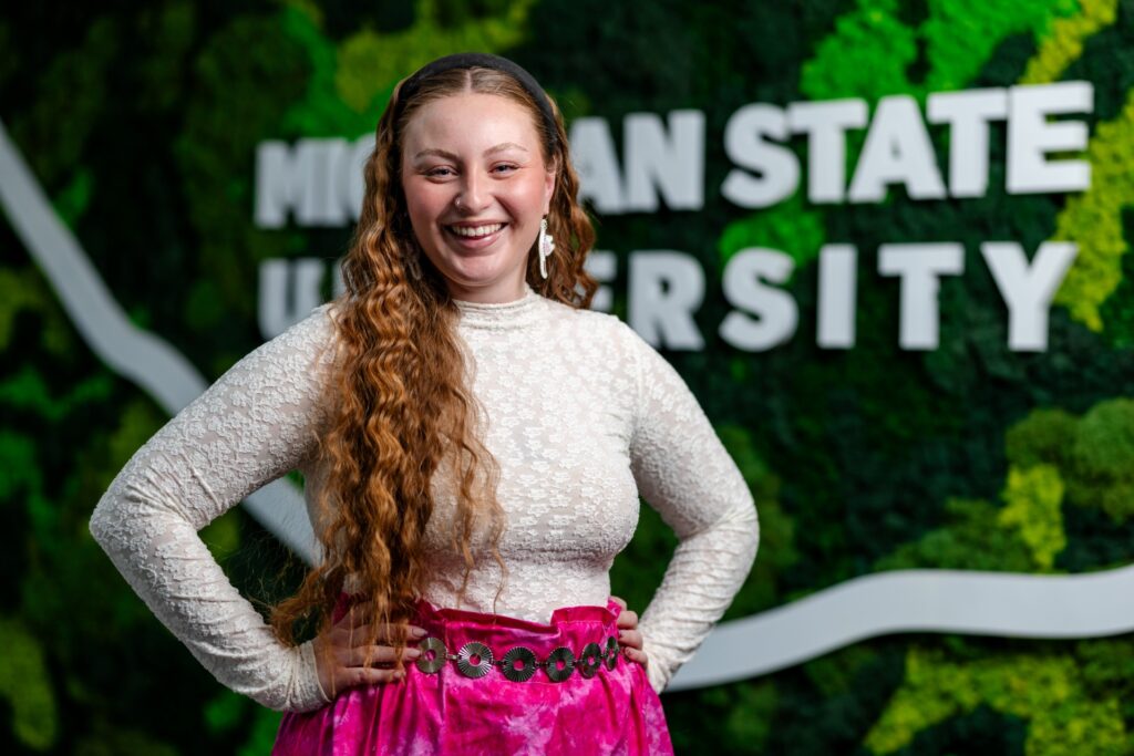 Smiling person with long wavy hair stands with hands on hips in front of a green wall display reading “Michigan State University,” wearing a light-colored lace top and a bright pink skirt.