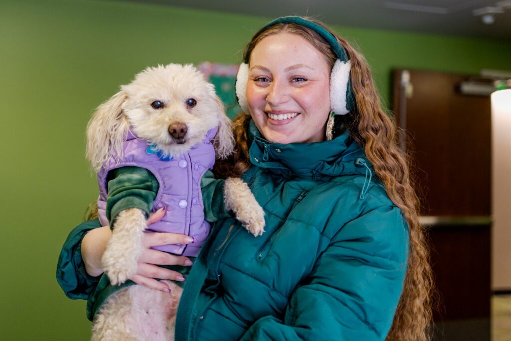 Smiling person wearing a teal winter coat and earmuffs holds a small white dog dressed in a purple vest and green sweater inside a building with green walls.