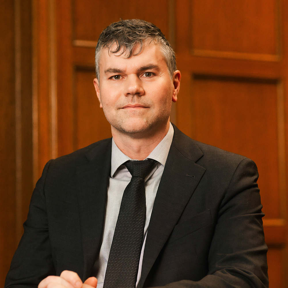 A man in a dark suit and tie sits against a wooden-paneled backdrop.