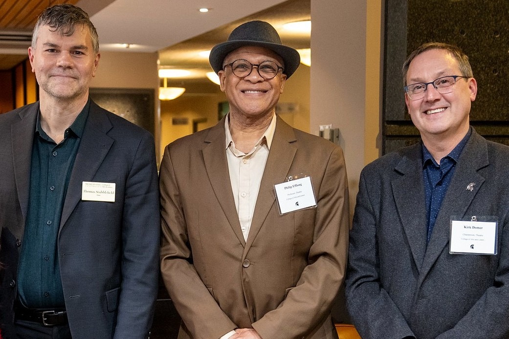 A medium shot of three men standing side-by-side at a formal university event. In the center, Philip Effiong, Professor of Theatre, wears a brown suit jacket, a fedora, and a smile. He is flanked by two colleagues in dark blazers; all three wear white MSU name tags. The background shows a warmly lit indoor space.