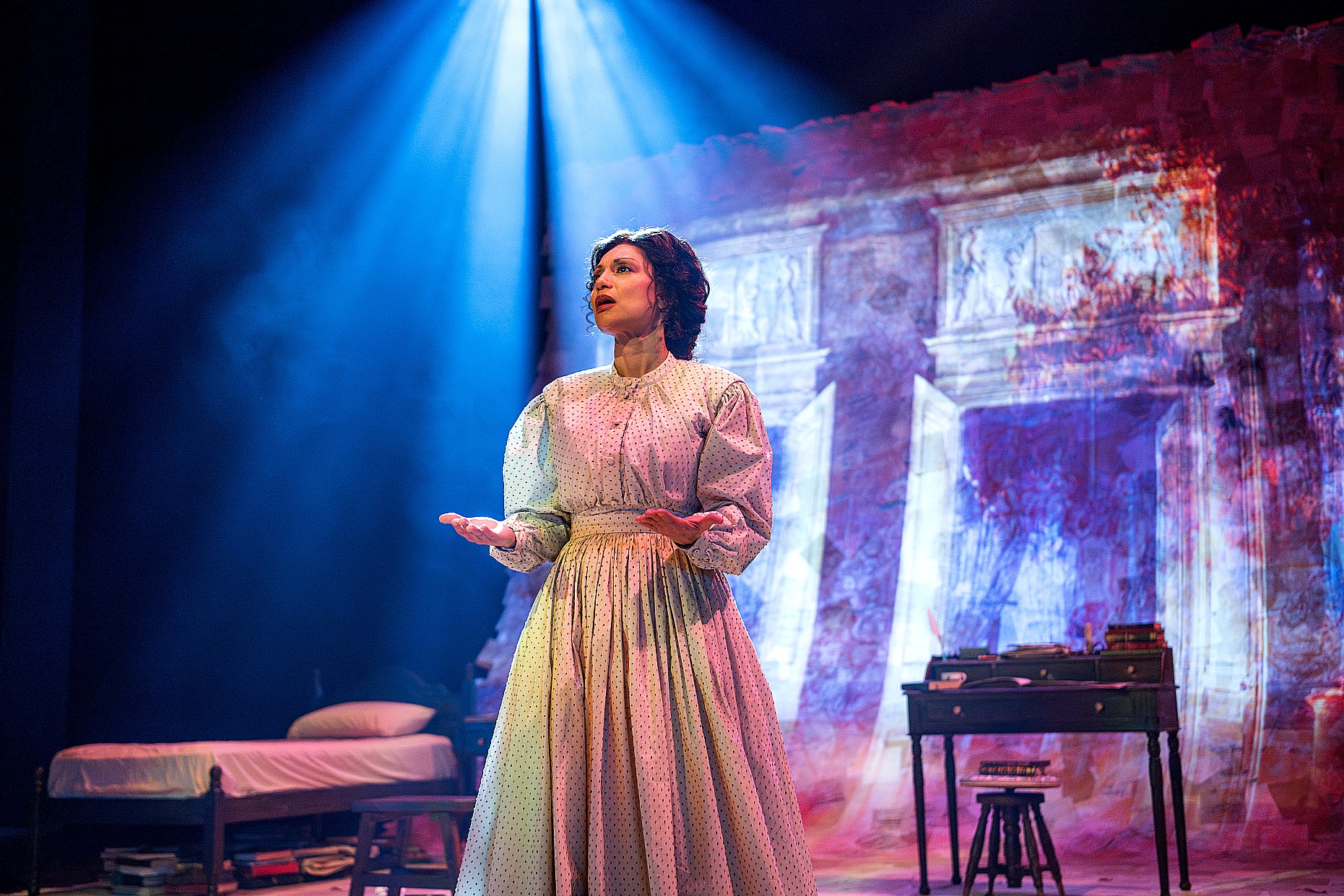 A woman in a cream period dress stands under a vibrant blue spotlight center stage with her palms turned upward. To her right is a wooden desk with a small lamp, and to her left is a bed. The background shows a digital projection of historical ledger lines and text.