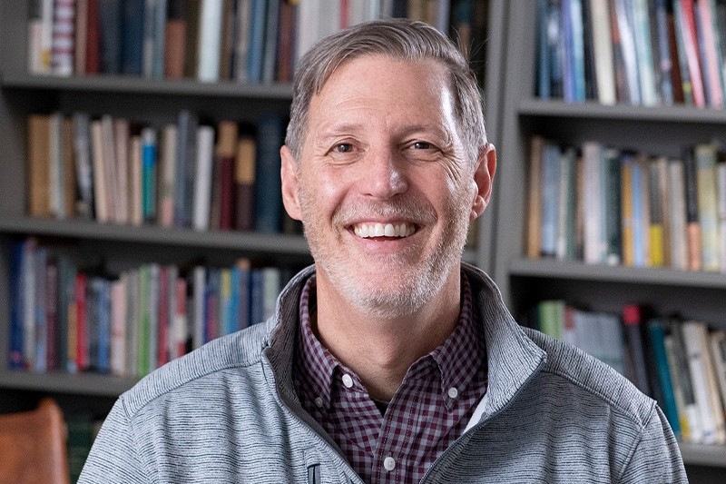 “Smiling man seated in an office or library setting, with bookshelves in the background, wearing a gray zip-up jacket.”