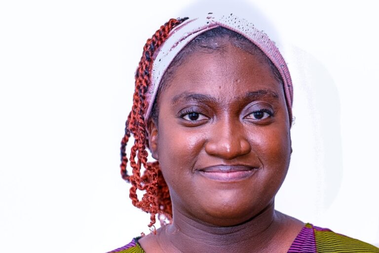 Portrait of a woman facing the camera against a white background, wearing a light headband, with braided hair pulled back and a slight smile.
