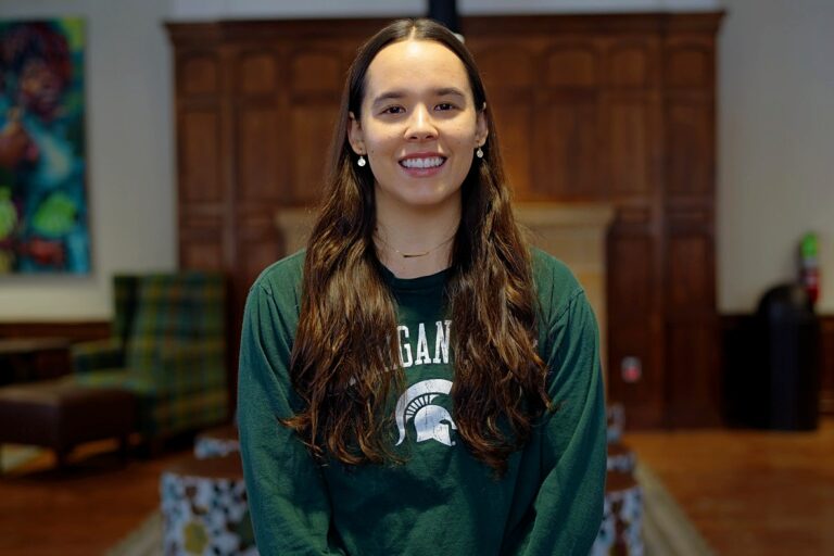Woman with long brown hair who is smiling and wearing a green Michigan State long-slevee T-shirt.