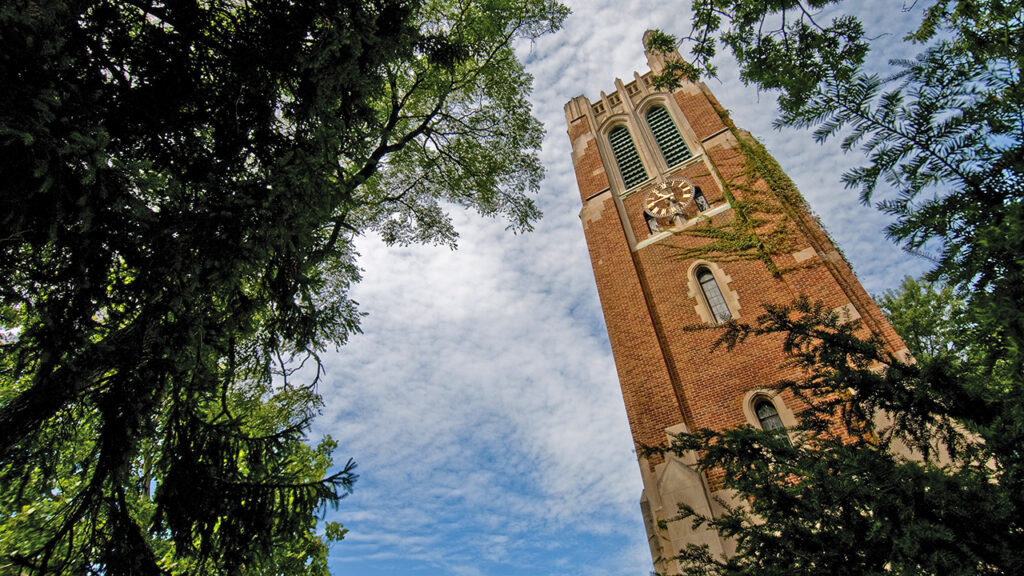 A low-angle view of MSU’s Beaumont Tower framed by lush green trees against a bright blue sky with wispy clouds