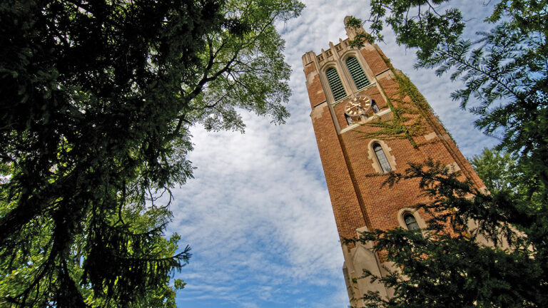 A low-angle view of MSU’s Beaumont Tower framed by lush green trees against a bright blue sky with wispy clouds