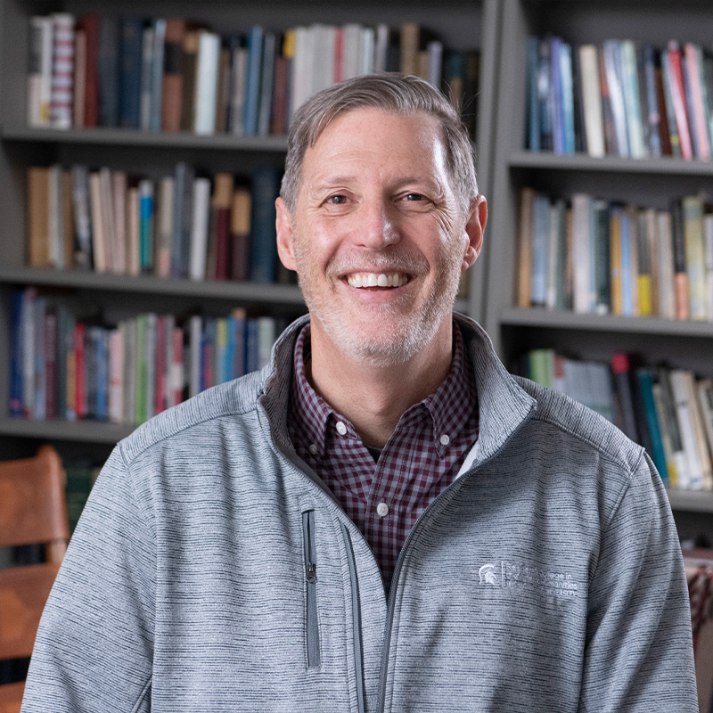 “Smiling man seated in an office or library setting, with bookshelves in the background, wearing a gray zip-up jacket.”