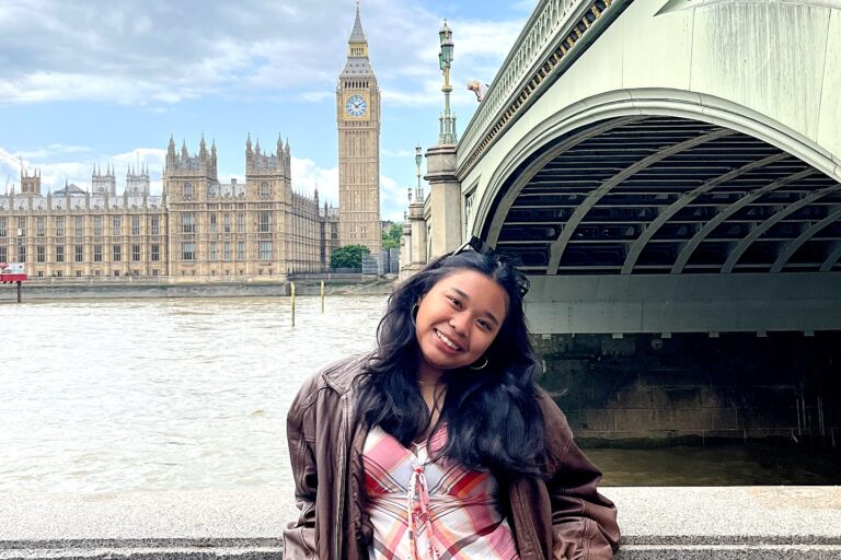 A young woman with long dark hair smiles while leaning against a stone railing in London. She wears a brown leather jacket over a plaid top and denim skirt. In the background, the River Thames flows past the Palace of Westminster and Big Ben, under a cloudy sky.