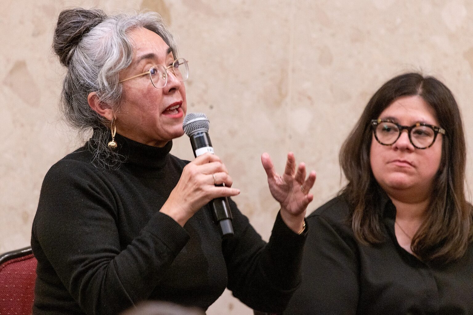 Cristina Rivera Garza speaks passionately into a microphone while gesturing with her open hand during the faculty panel discussion, as Alejandra Márquez Guajardo — wearing dark-framed glasses and a black top — listens attentively beside her.