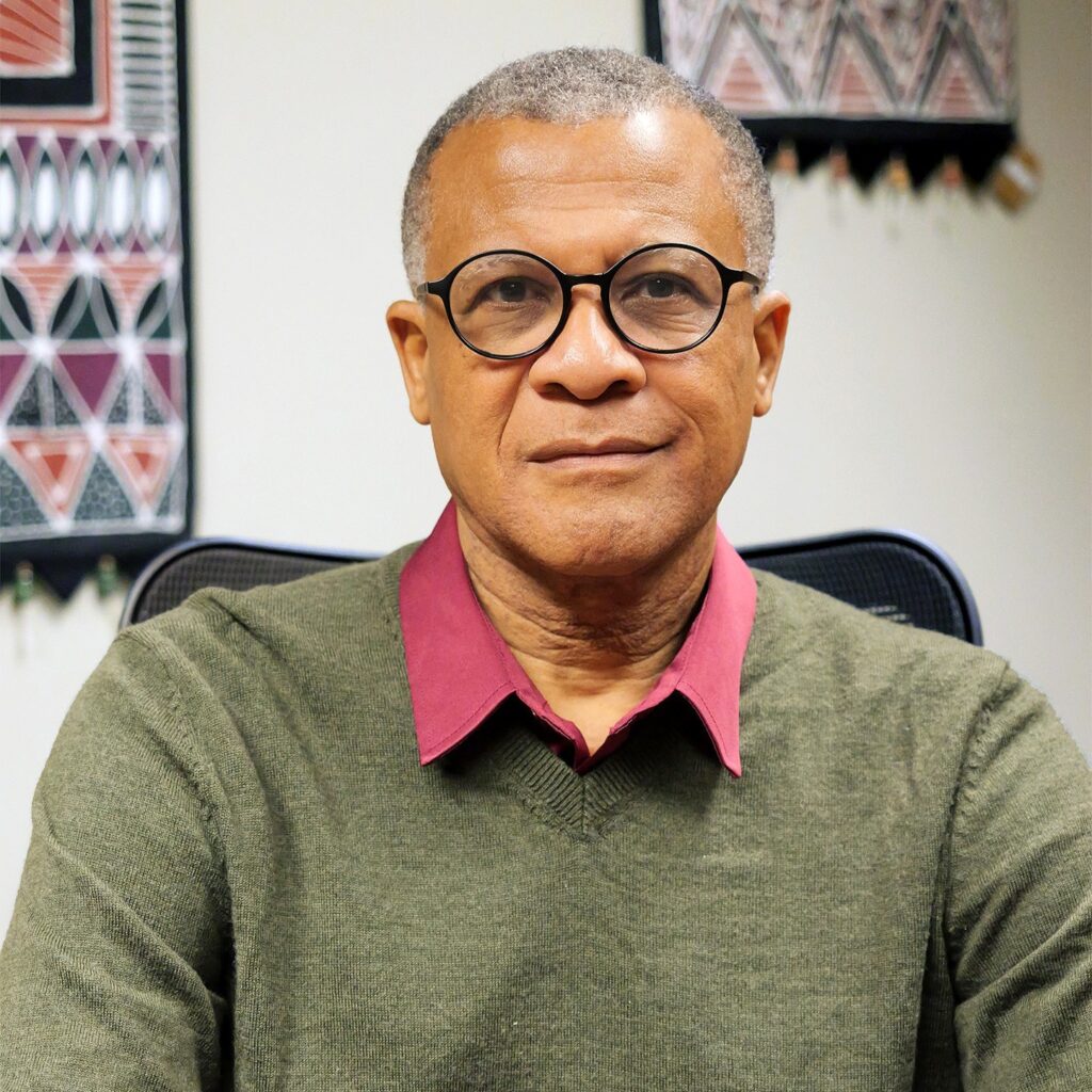 A headshot of Philip Effiong, a man with short graying hair, looking directly at the camera with a subtle smile. He is wearing distinctive round black glasses, an olive green v-neck sweater, and a maroon collared shirt. He is seated in an office with framed traditional textile art featuring geometric patterns hanging on the white wall behind him.