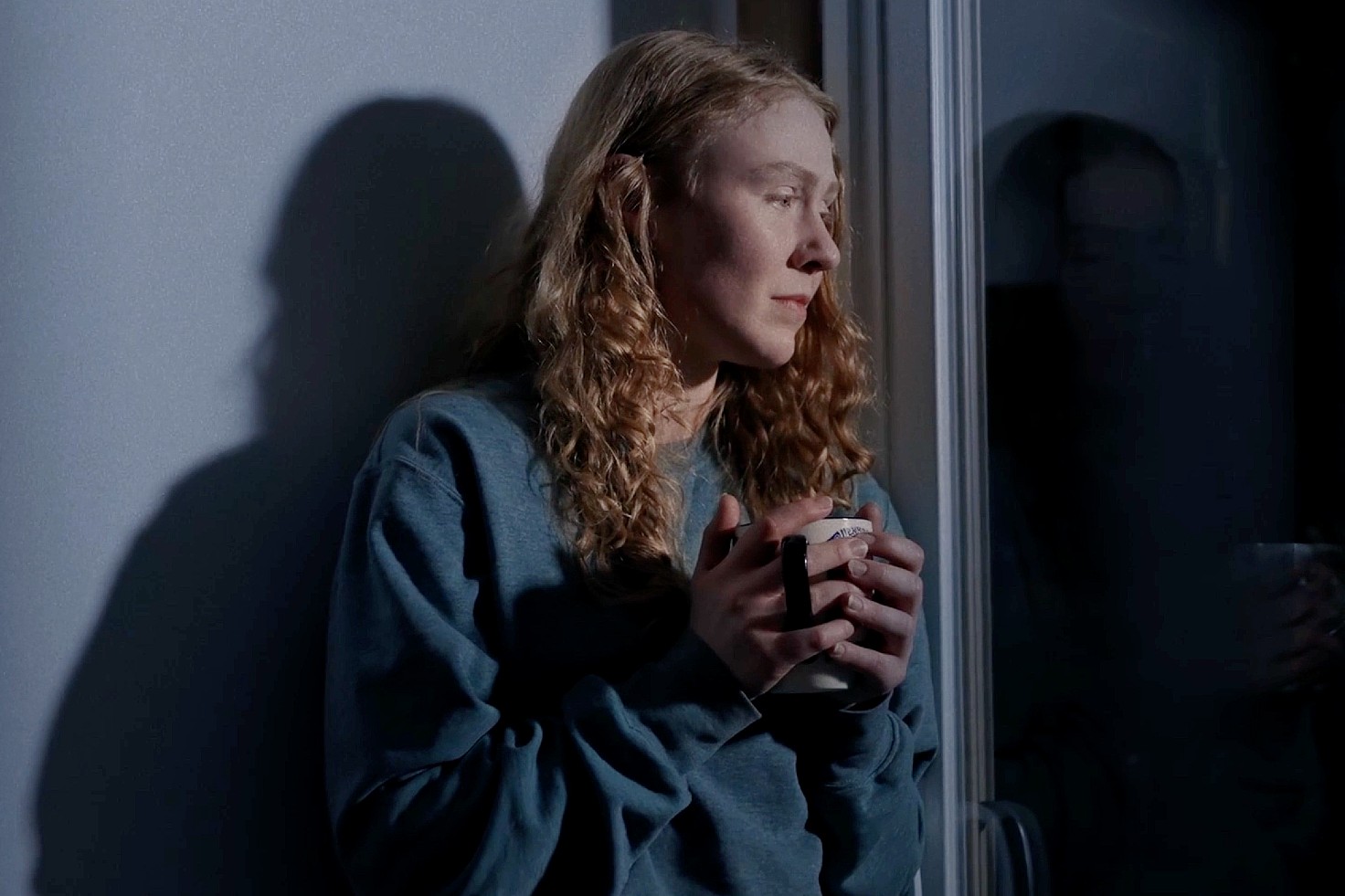 A person with long hair holds a mug, gazing thoughtfully out a window. They're wearing a blue sweater, and their reflection is visible, creating a contemplative mood.