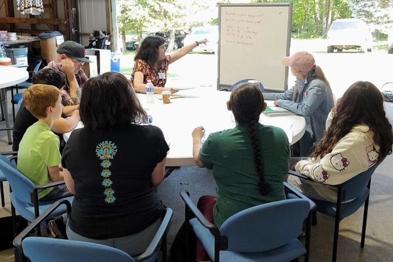 Children and adults seated in blue chairs around white tables in an outdoor pavilion. A woman in burgundy clothing points at a whiteboard. Participants include a child in a yellow-green shirt, a person in a black shirt with teal design, and a person in a tan/cream colored blanket. Sunlight and green outdoor landscape visible through the open structure.