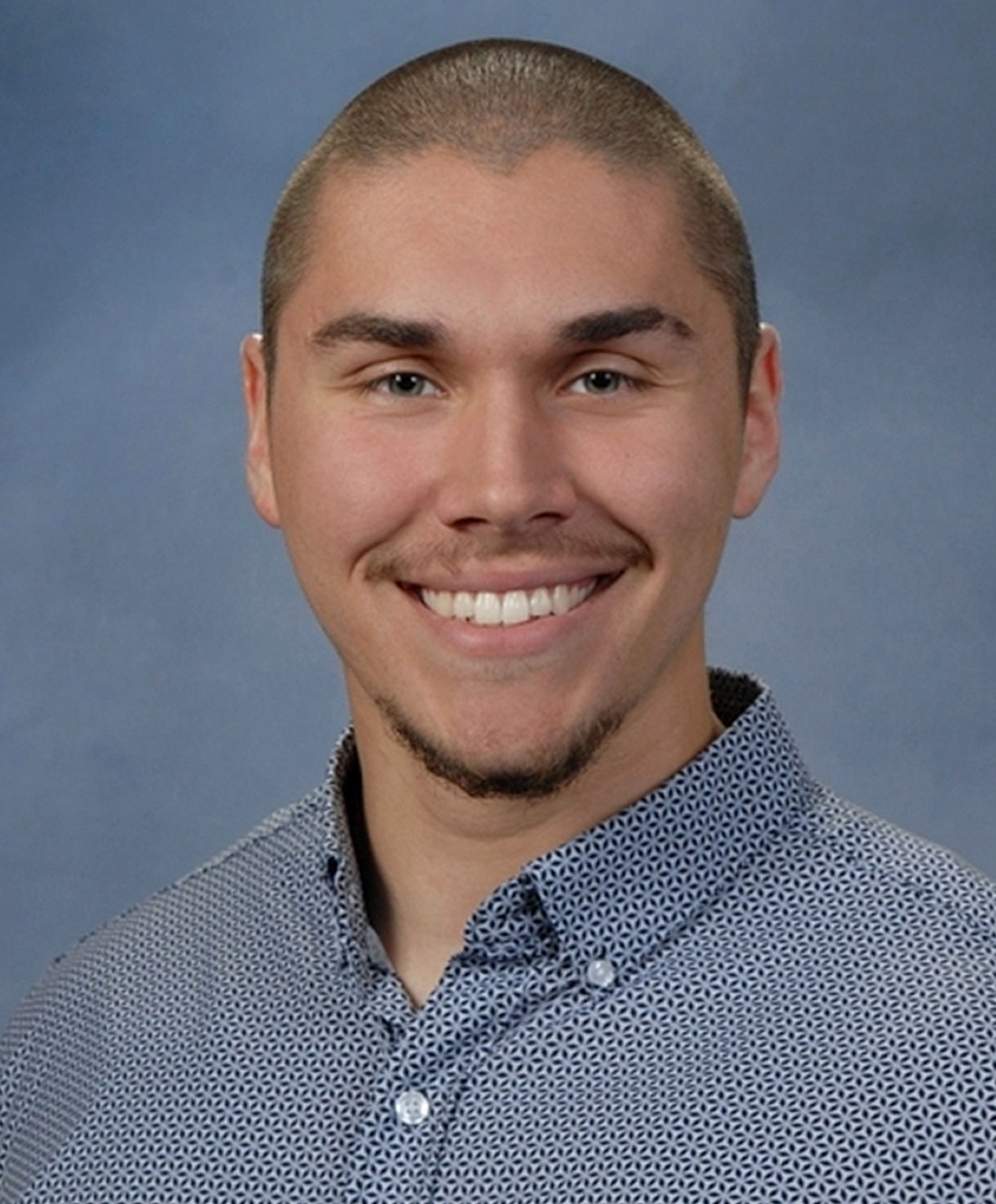 Smiling young adult male with short hair, wearing a patterned button-up shirt against a soft blue background. Mood is friendly and welcoming.