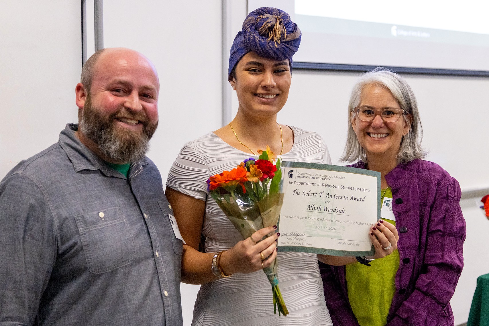 Three people smiling; the center person holds flowers and an award certificate. The atmosphere is celebratory, suggesting an awards ceremony.