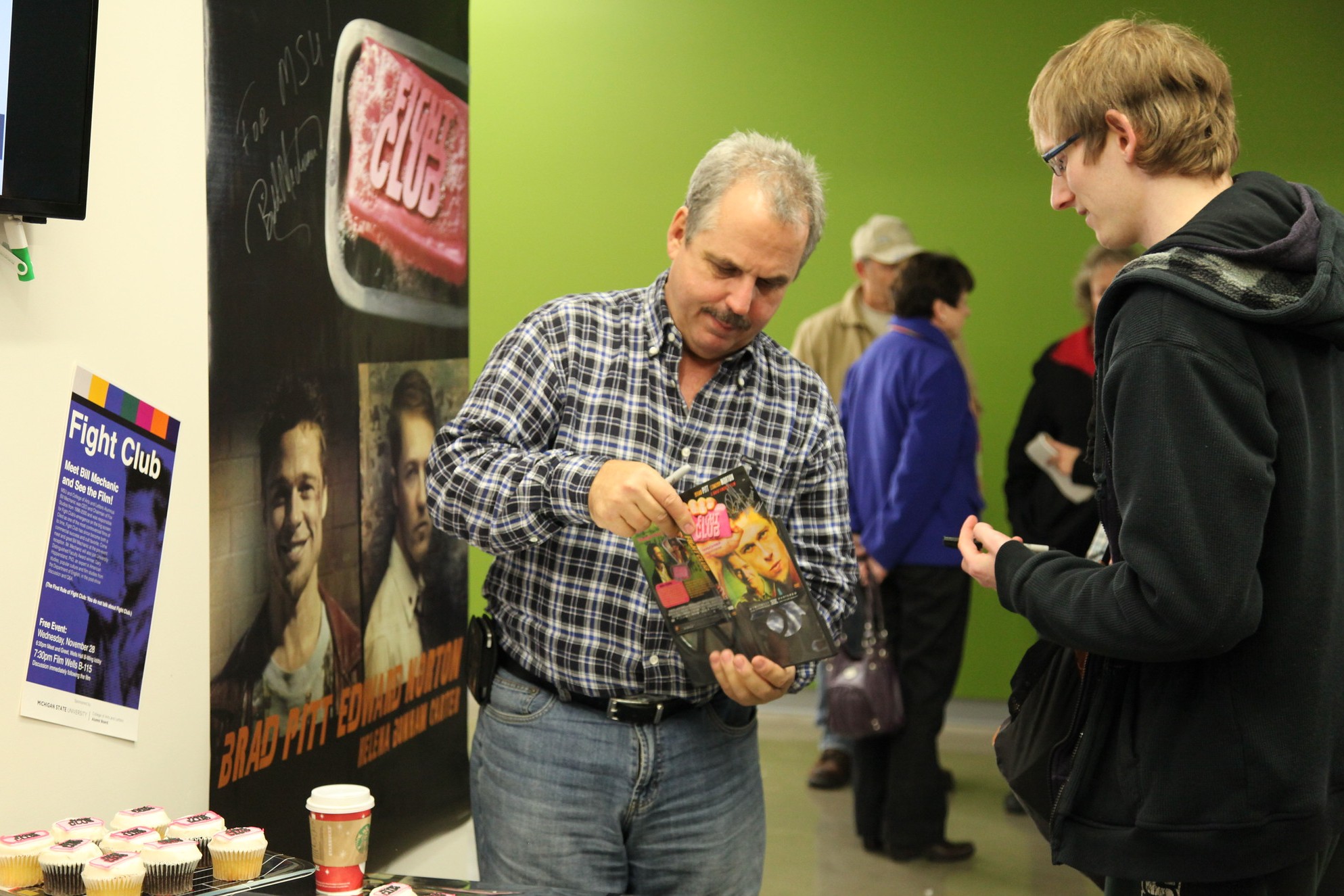 A man in a plaid shirt signs a "Fight Club" book for a young man in glasses. A poster and cupcakes are on a table, with a green wall in the background.
