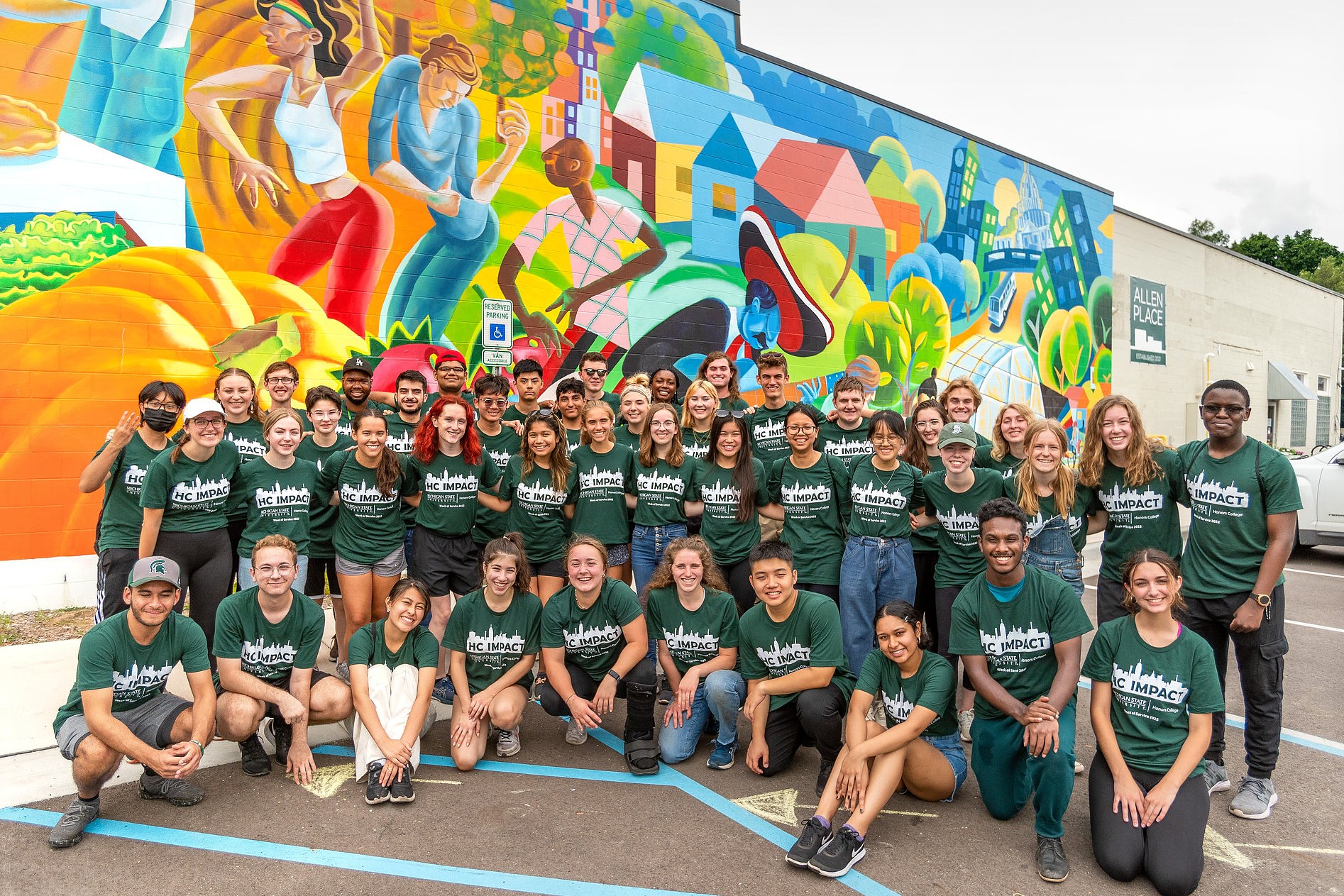 A large group of smiling people wearing matching green shirts poses in front of a vibrant mural with abstract figures and colorful houses. The scene conveys a sense of unity and community involvement.