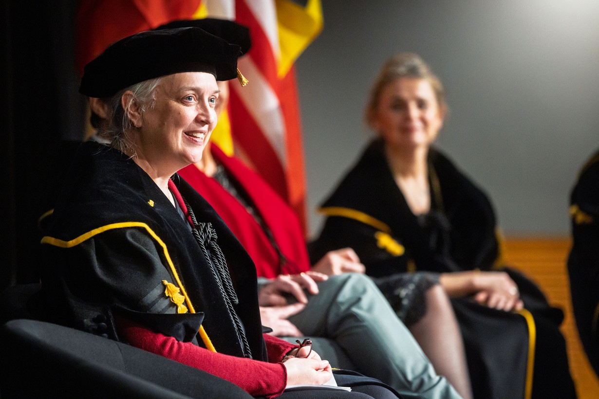 A woman in academic regalia smiles while seated onstage at a formal event. Behind her are U.S. flags and more seated individuals, suggesting a graduation or ceremony.
