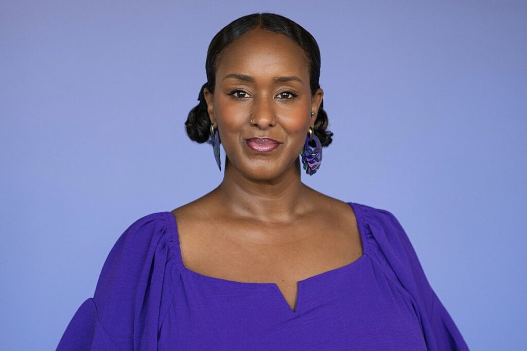 A woman in a vibrant purple dress stands against a lavender background. She wears matching earrings, and her expression is confident and serene.