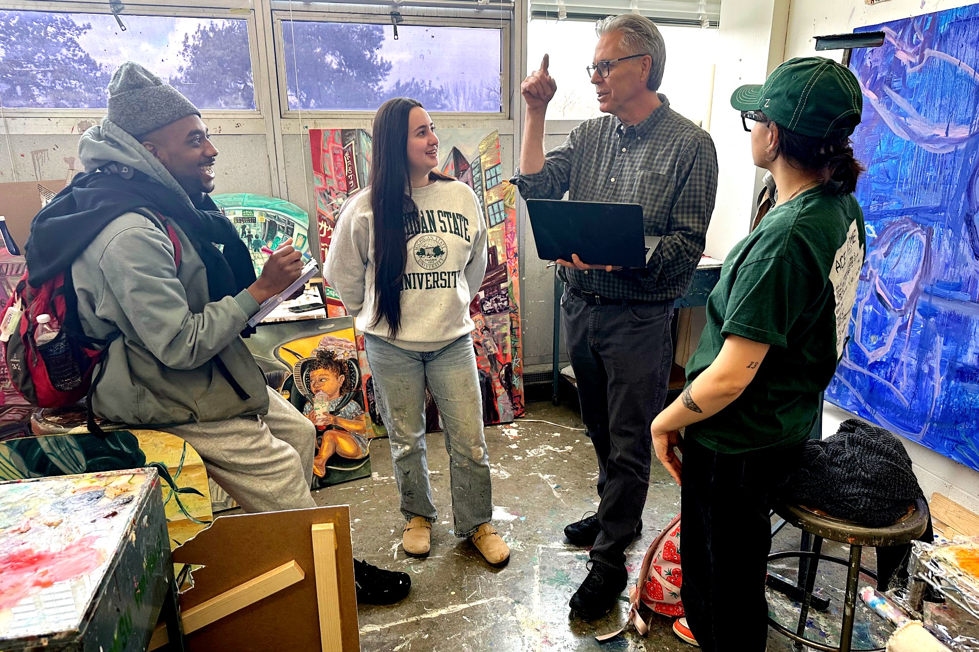 A group of four people in an art studio engage in a lively discussion. The room is filled with vibrant paintings, creating a creative and energetic atmosphere.