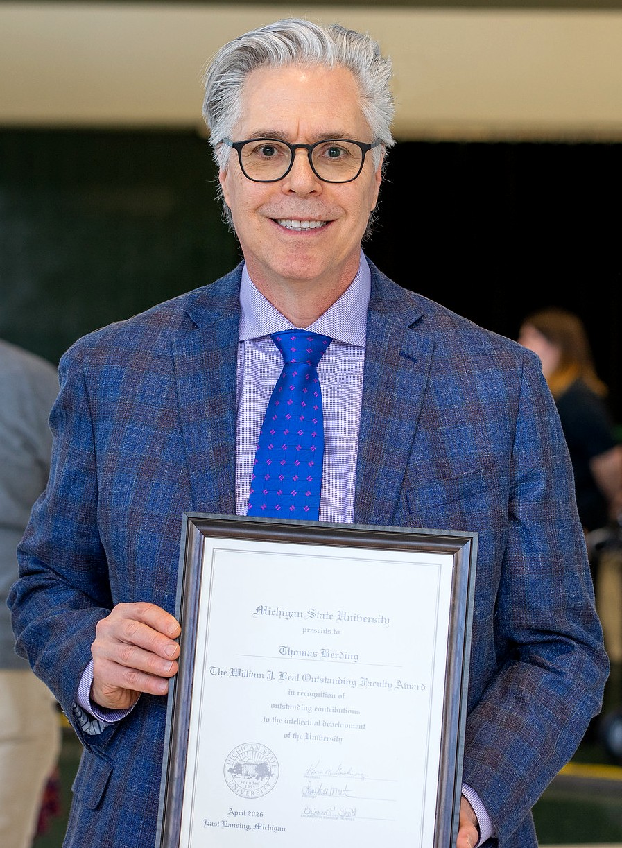 Man in glasses and a blue plaid suit holds a framed award. He smiles proudly in a professional setting, conveying accomplishment and joy.