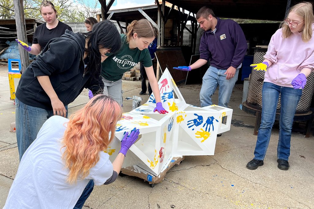 A group of people is painting handprints in colorful paints on a white, sculptural object outdoors. They wear gloves and appear focused and collaborative.