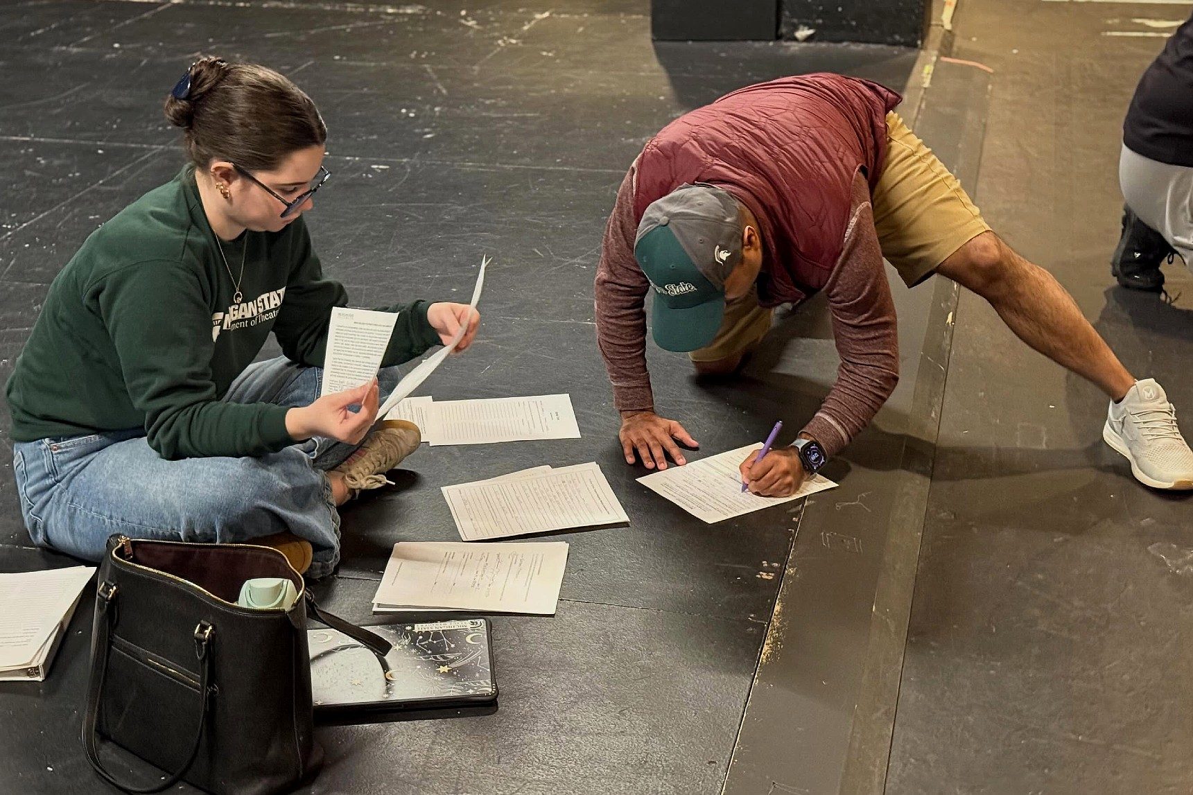 Two people sit and stretch on a theatre stage floor while reviewing papers during what appears to be a rehearsal or workshop. One person sits cross-legged reading documents, while another in Michigan State gear stretches in a lunge position and writes on a sheet of paper surrounded by scripts and materials.